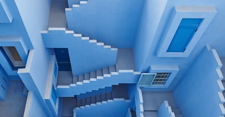 AI conflicting objectives: interlocking blue staircases in a geometric facade, each flight leading somewhere different